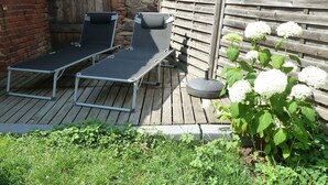 Een houten terras met twee lounge fauteuils en een kleine tafel, omringd door een bakstenen muur en een houten hek.