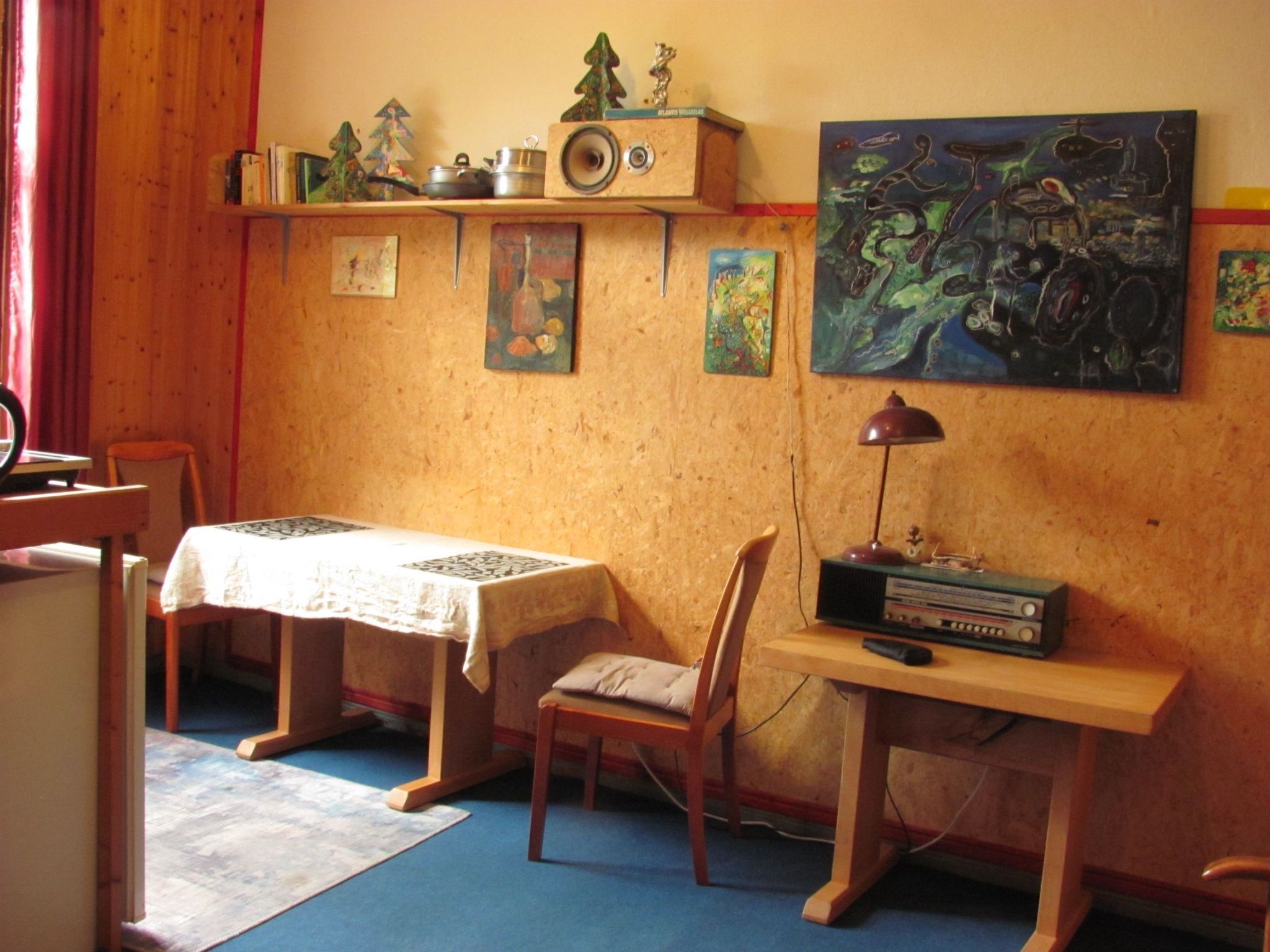 Una habitación con dos mesas de madera, una con mantel blanco y otra con un radio. Hay sillas, una estantería con objetos decorativos y pinturas en la pared.