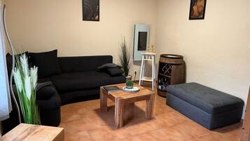 Ein Wohnzimmer mit einer schwarzen Couch, einem Holztisch, einem grauen Hocker und einem kleinen weißen Tisch mit Dekorationen.