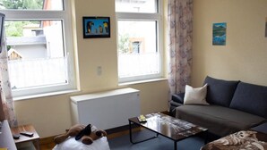 Una sala de estar con un sofá, una mesa de centro y un sillón de peluche. Hay ventanas con cortinas, un radiador y un cuadro colgado en la pared.