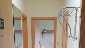 Ein schmaler Flur mit Holzboden führt zu einem Schlafzimmer mit Etagenbett. Eine Bank steht an der Wand, und ein Metallregal ist an der Wand befestigt.