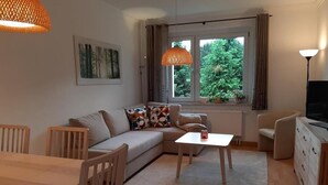 Ein modernes Wohnzimmer mit einer Couch, einem Esstisch und Stühlen. Ein Fenster mit Vorhängen, ein Bild an der Wand und eine Lampe sind ebenfalls zu sehen.