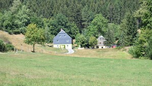 Un paysage rural avec un grand champ, une maison au toit en pente et une colline boisée en arrière-plan.