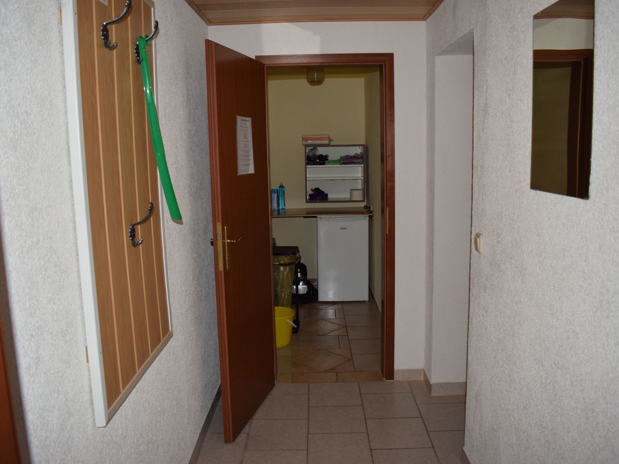 Un pasillo estrecho con una puerta de madera a la izquierda, una toalla verde colgada de un perchero, y una pequeña zona de kitchenette con un refrigerador y un lavabo al fondo.