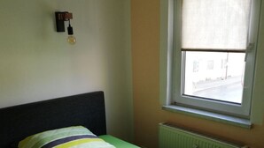 Un lit simple avec un matelas vert et une couverture multicolore, dans une pièce dotée d’une fenêtre et d’un radiateur.