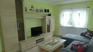 Una sala de estar con un sofá, un televisor sobre un mueble de madera, una mesa de centro de vidrio y una ventana con cortinas.