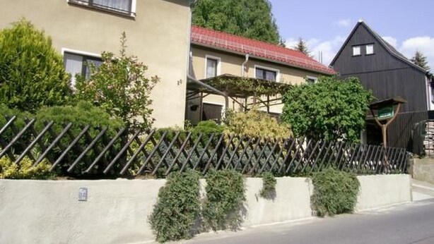 Un edificio residencial con techo de tejas rojas, un porche cubierto y un patio cercado con vegetación.