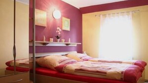Un dormitorio con cama, cómoda, espejo, estante con objetos decorativos y ventana con cortinas.