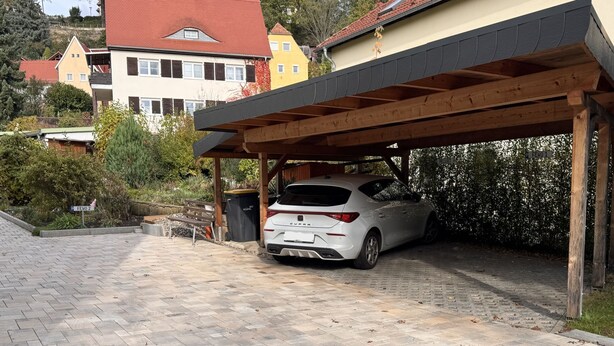 Un carport coperto con un'auto bianca parcheggiata all'interno, un vialetto lastricato e case sullo sfondo.