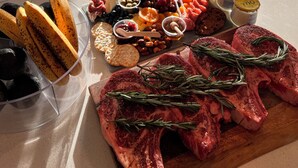 Une table à manger avec une planche de charcuterie et de la viande crue, un canapé et une fenêtre donnant sur des immeubles.