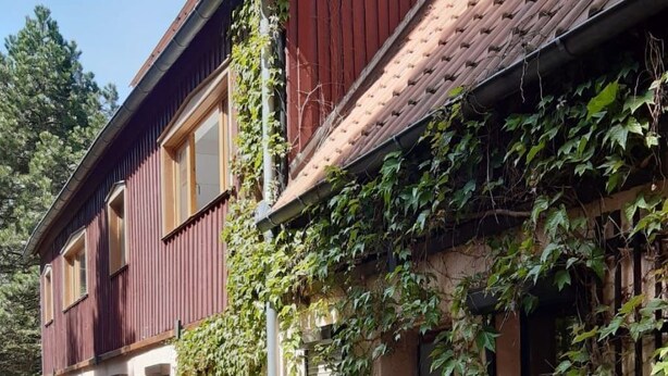 Een twee verdiepingen tellend huis met een rode gevel en een pannendak. Een verharde oprit bevindt zich ervoor, en het huis wordt omringd door groen en planten.