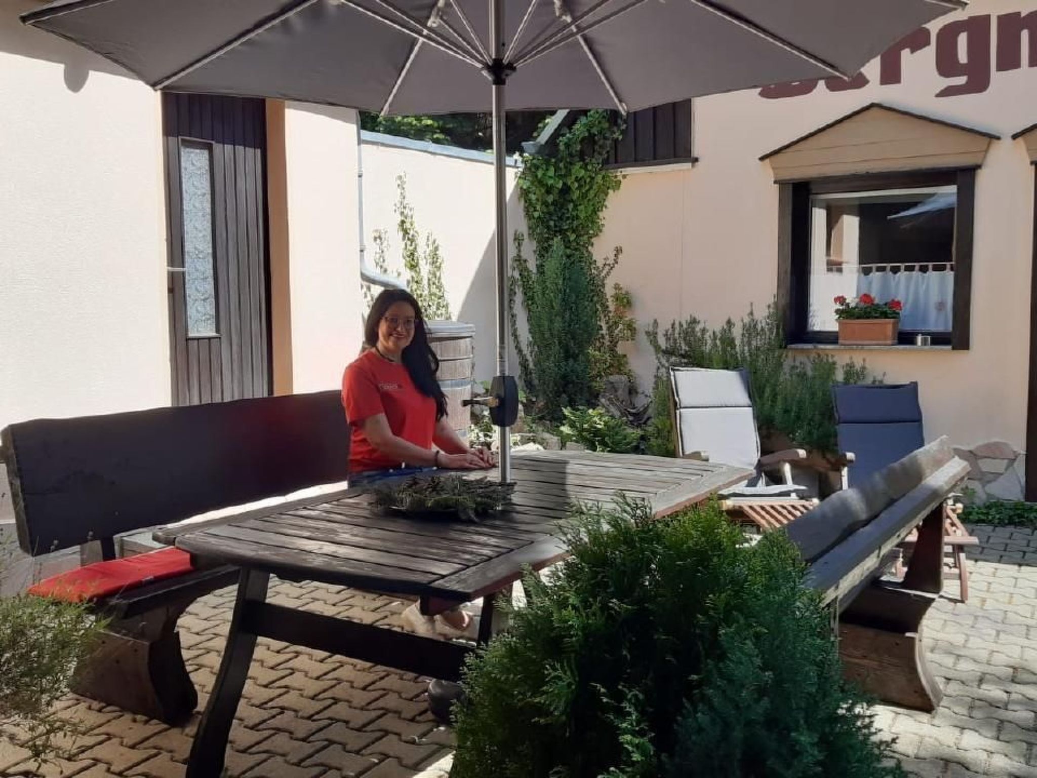 An outdoor patio with a wooden table and benches, surrounded by potted plants and a building with a sign.