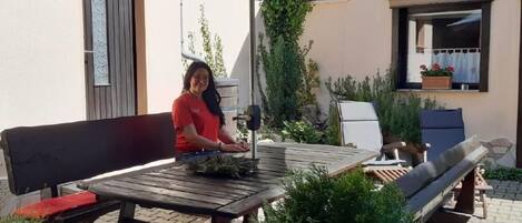 An outdoor patio with a wooden table and benches, surrounded by potted plants and a building with a sign.