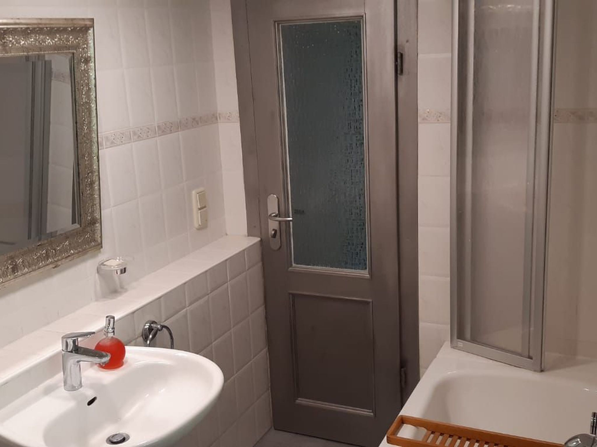 Un baño con un lavamanos blanco, un espejo con un marco ornamentado, una puerta con panel de vidrio esmerilado y una zona de ducha.
