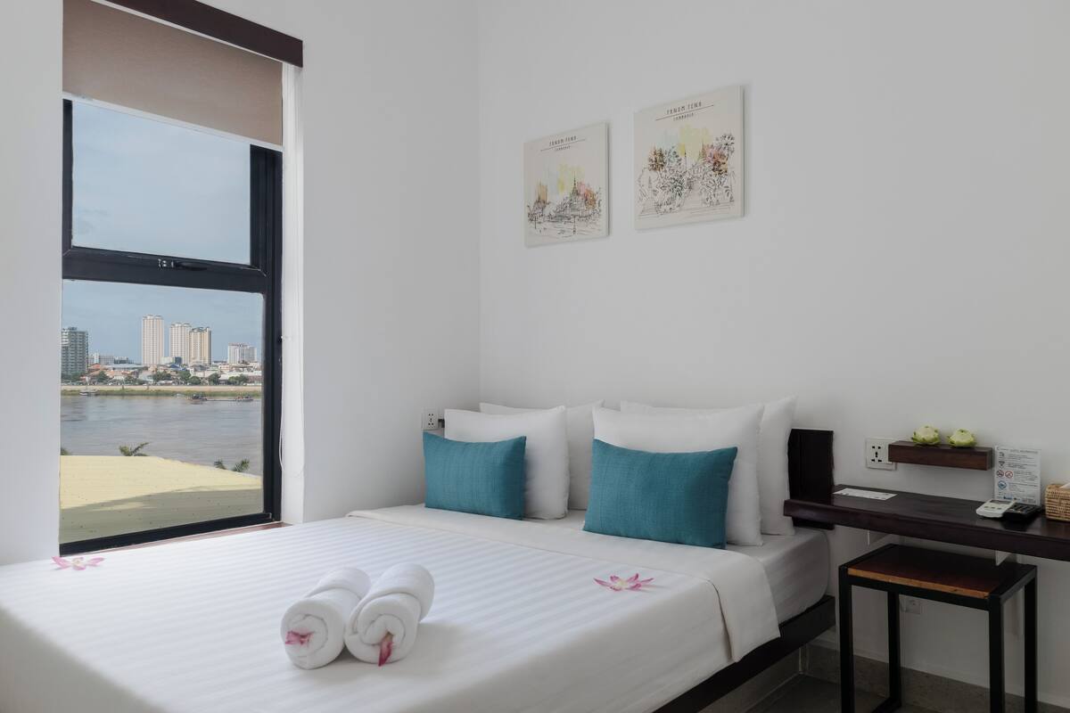 A hotel room with a bed, a desk, and a view of a cityscape through a large window.
