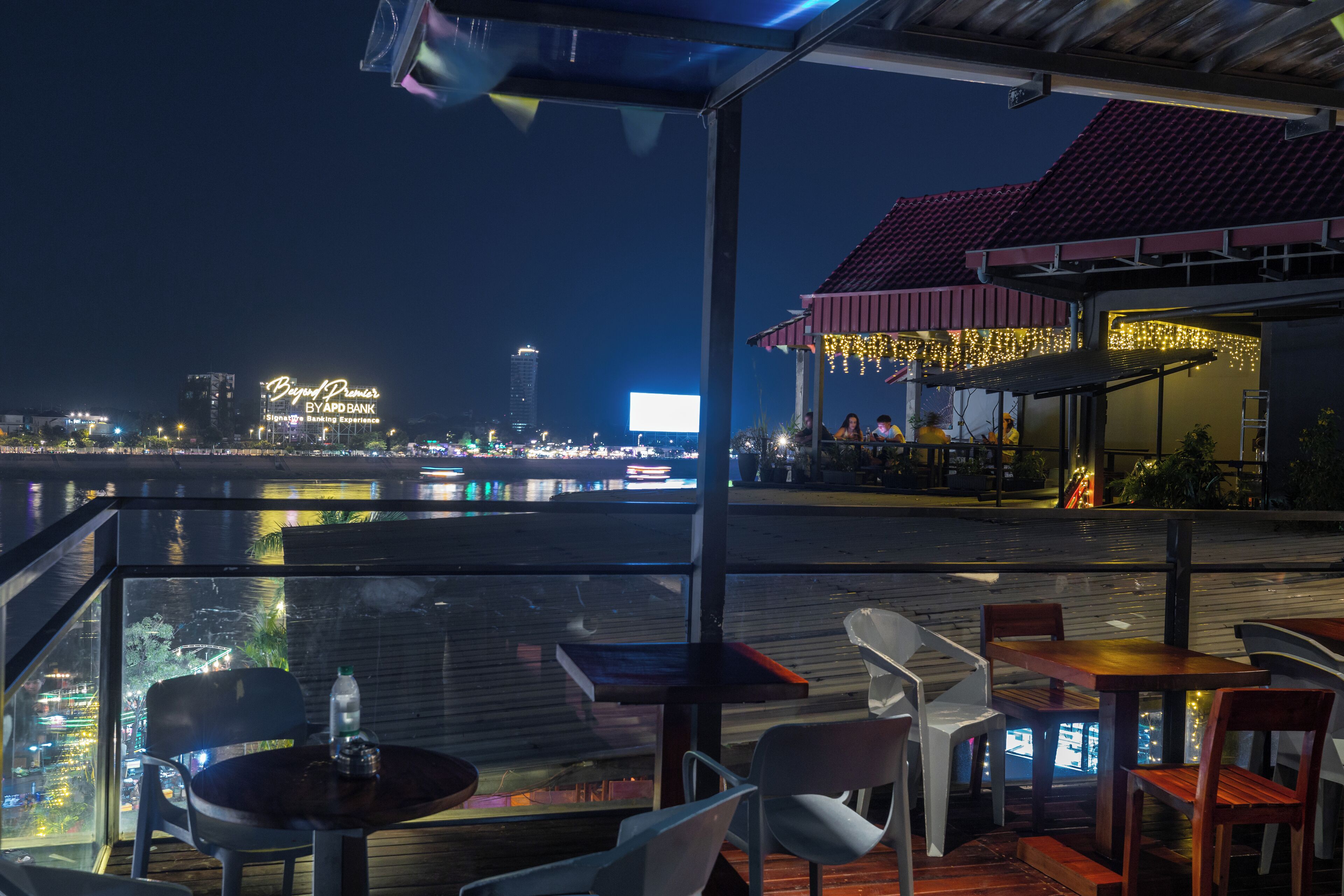 An outdoor dining area with tables and chairs, overlooking a waterfront with city lights and a large billboard in the background.
