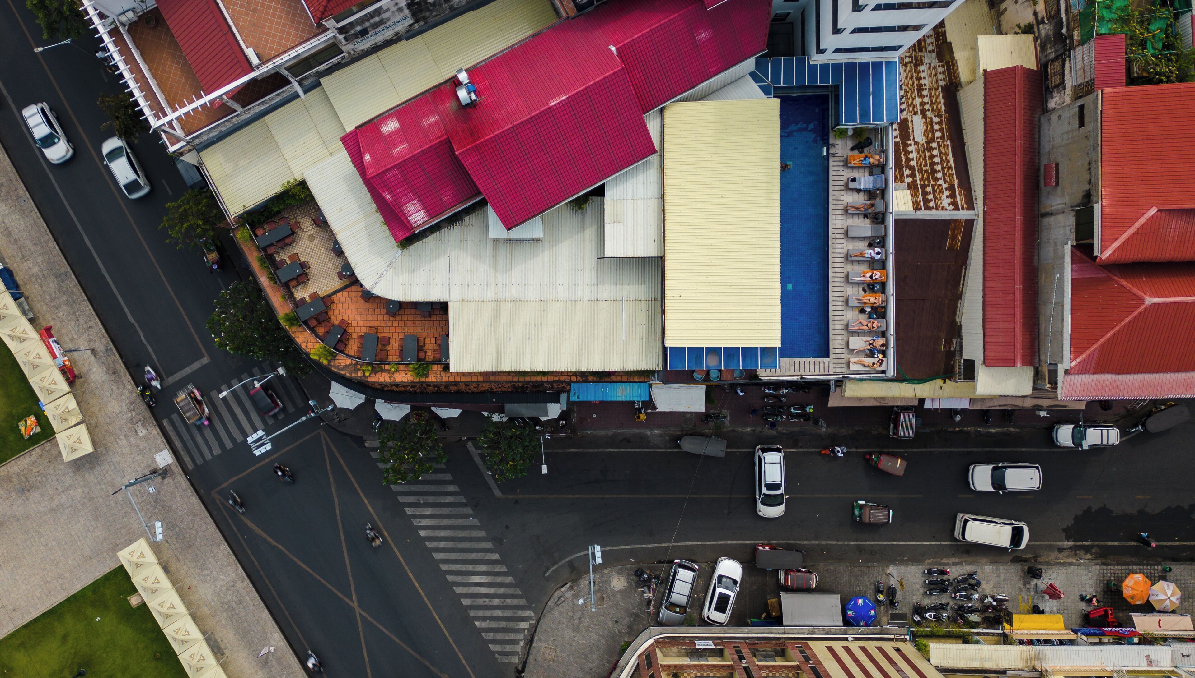 An aerial view of a building with a red roof, a blue swimming pool, and a street with cars and a pedestrian crossing.