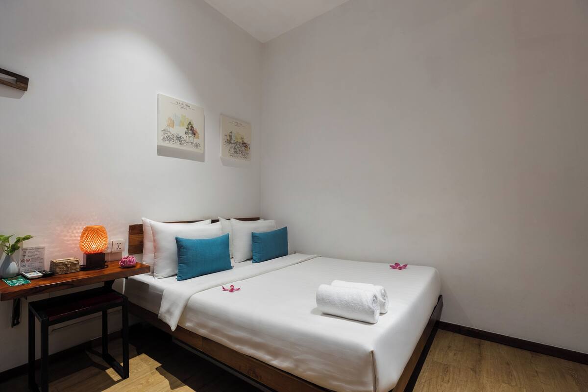 A neatly made bed with white linens, blue pillows, and a rolled towel. A small table with a lamp and a plant is beside the bed.