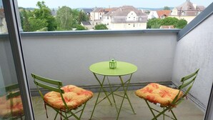 Un balcon avec deux chaises vertes et une petite table ronde, une lanterne verte, et une vue sur des maisons et des arbres en arrière-plan.