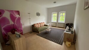 Un salon spacieux comprenant un canapé, une table basse et un tapis. Il y a deux fenêtres équipées de stores, une lampe sur pied et une petite table avec un ordinateur.