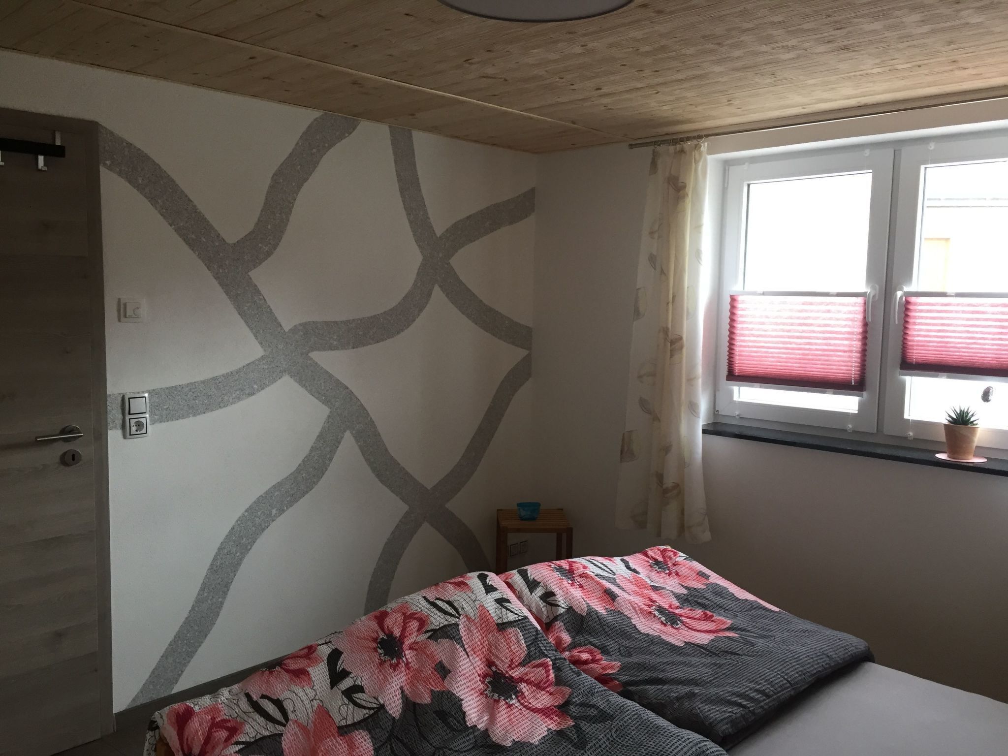 A bedroom with a bed, a window with red blinds, a small plant on the windowsill, and a wall with a geometric pattern.