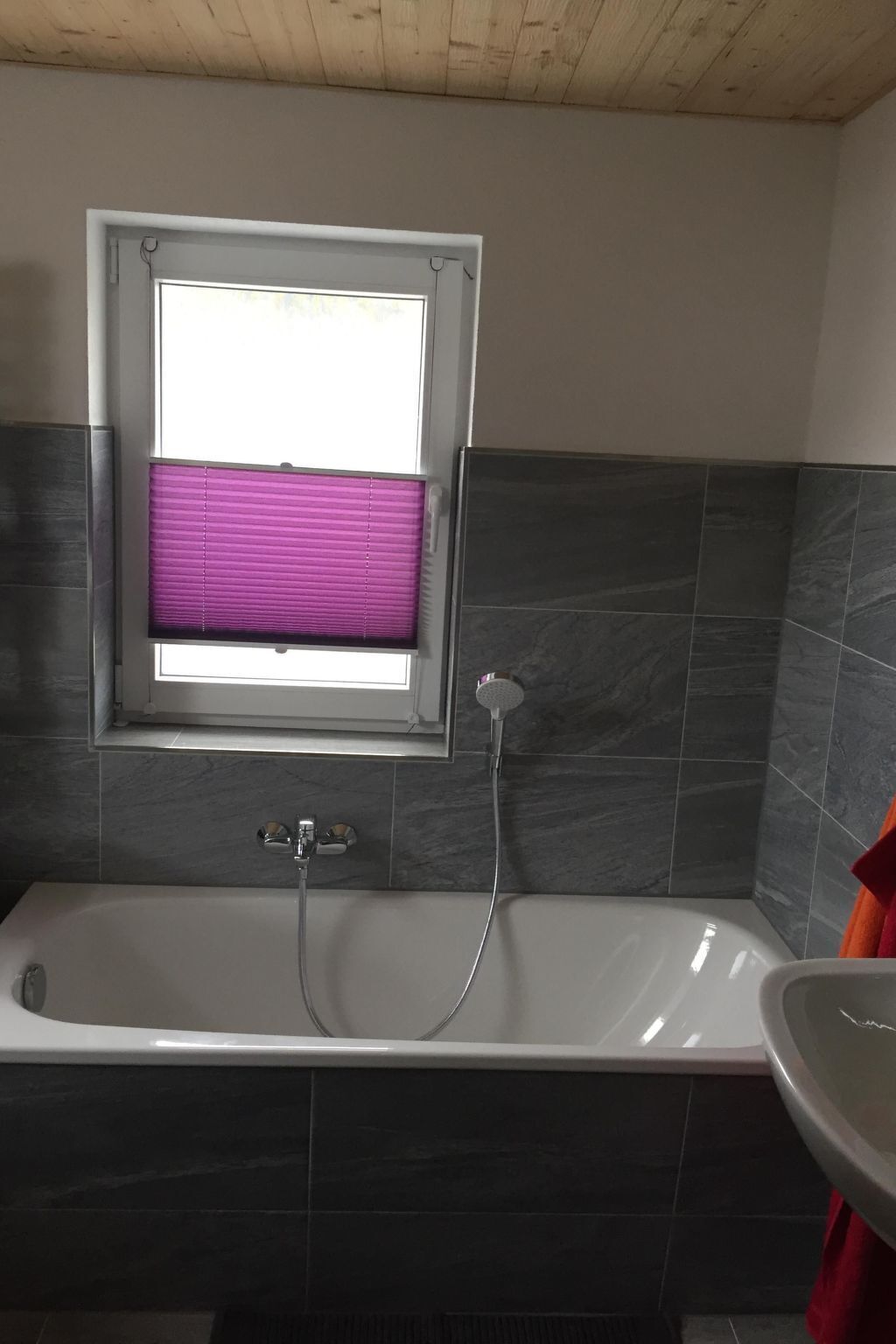 Ein Badezimmer mit einer Badewanne, einem Fenster mit violetten Rollos und dunkel gefliesten Wänden.