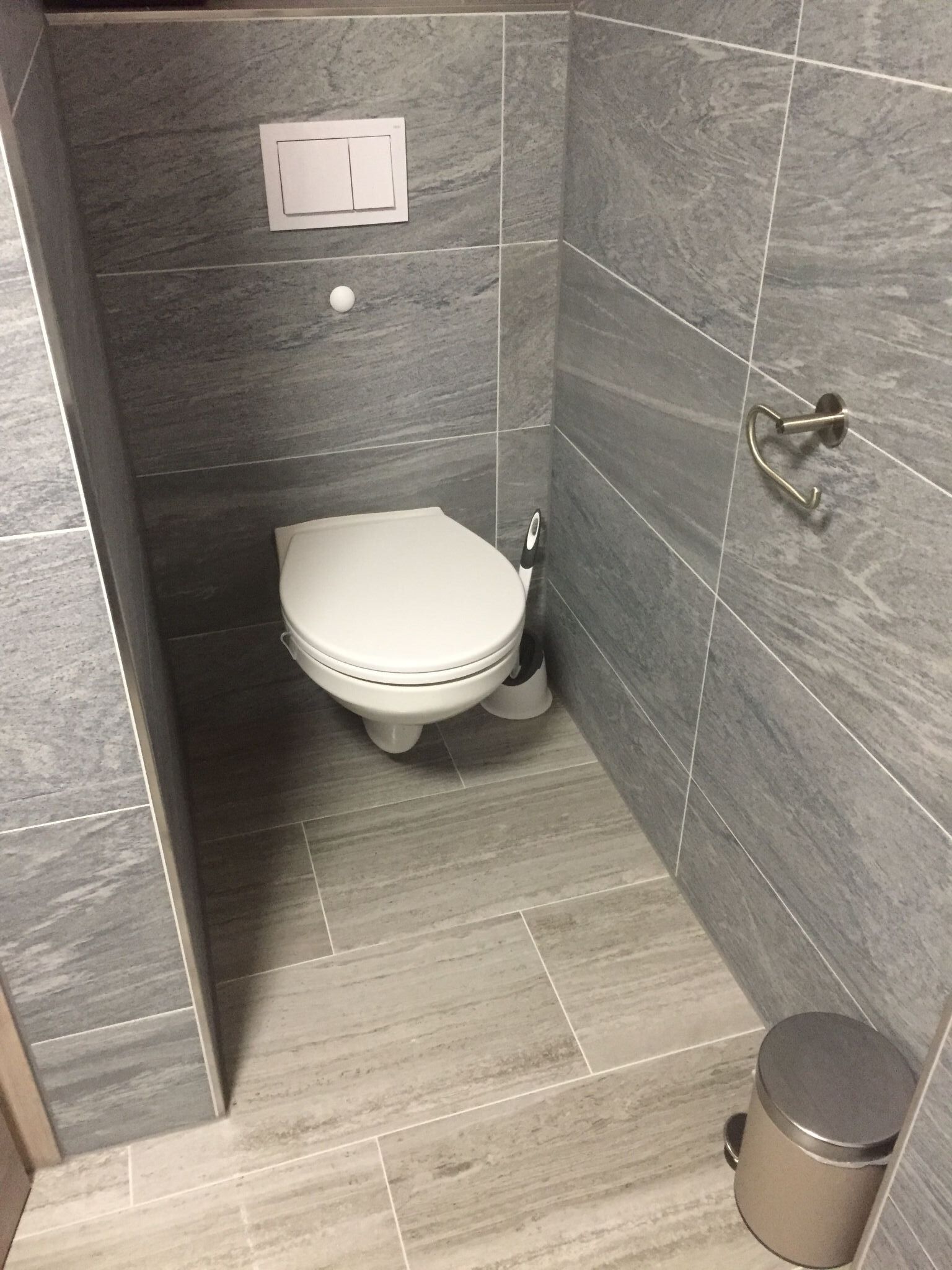 A small, modern bathroom with a toilet, a wall-mounted flush handle, and a trash can.