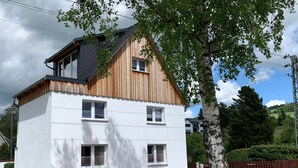 Une maison à deux étages, avec une façade blanche et une partie supérieure en bois, dotée de trois fenêtres et d’une cheminée visible.
