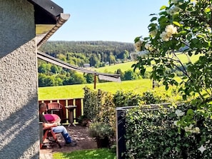Eine Terrasse mit Blick auf ein grünes Feld und Bäume, eine Person liest und eine Hängematte ist aufgehängt.