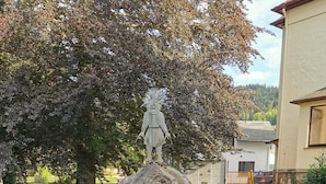 Eine Statue auf einem Felsen mit einer Gedenktafel, umgeben von grünen Büschen, mit einem Baum und einem Gebäude im Hintergrund.