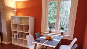Ein Essbereich mit einem Tisch für vier Personen, umgeben von weißen Stühlen, einem Bücherregal und einem Fenster mit Blick auf Bäume.