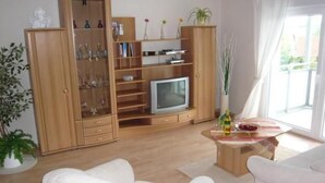 A living room with a wooden cabinet, a television, a white sofa, a small table with a bottle, and a window with curtains. - in Witzelroda mit Grill und Garten by Interhome (Moorgrund)