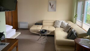 A living room with a beige sofa, a small round table, a flat-screen TV, and a window with a view of greenery.