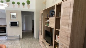 Una sala de estar moderna con un mueble de madera para entretenimiento, un televisor y una estantería. Se aprecia una cocina al fondo con estufa y armarios.