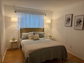 A neatly arranged bedroom with a bed, two bedside lamps, a small table with a plant, and two framed pictures on the wall.