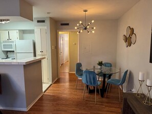A modern kitchen with white cabinets, a microwave, and a refrigerator. There is a dining area with a glass table and blue chairs. A unique chandelier hangs from the ceiling.