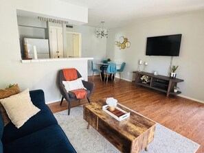 A living room with a dark blue sofa, a wooden coffee table, and a dining area with blue chairs. A TV is mounted on the wall, and there is a small table with candles and a plant.