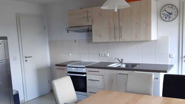 A compact kitchen with a stove, sink, and cabinets. There is a dining table with chairs and a clock on the wall. - Familienfreundliche Wohnung in Vorland mit Garten by Interhome (Grimmen)