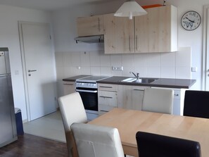 A compact kitchen with a stove, sink, and cabinets. There is a dining table with chairs and a clock on the wall. - Familienfreundliche Wohnung in Vorland mit Garten by Interhome (Grimmen)