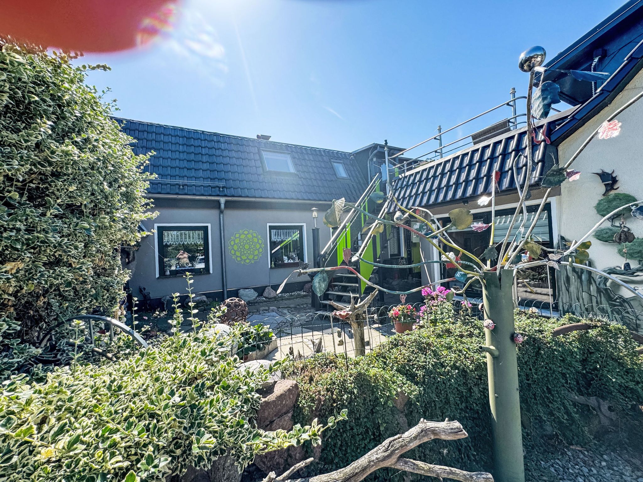 A residential area with a house featuring a tiled roof, a staircase, and a garden with various plants and a metal sculpture.