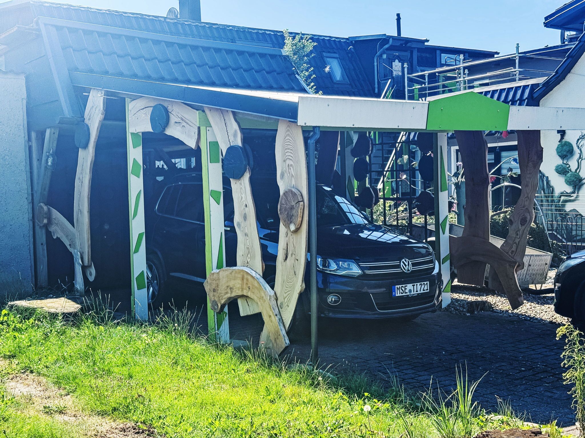 Ein Auto steht unter einer Holzbeschalung mit grüner Begrünung, umgeben von Gras und einem befestigten Bereich.