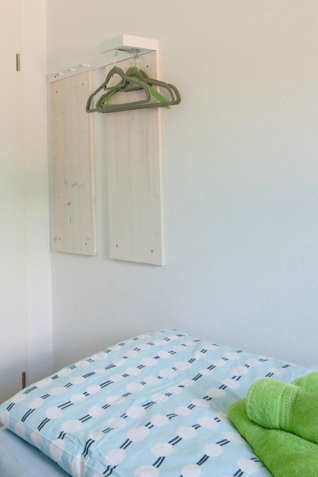 Ein Zimmer mit einem Bett, das eine hellblaue und weiße gepunktete Bettdecke, ein grünes Handtuch und einen an der Wand befestigten Wäscheständer mit zwei Kleiderbügeln zeigt.