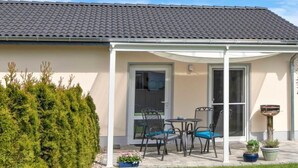 Ein Haus mit überdachter Terrasse, Rasen und hohem Heckenstreifen.