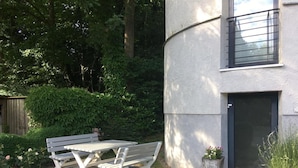 Un patio con una mesa y bancos, rodeado de vegetación y un edificio con una puerta y una ventana.