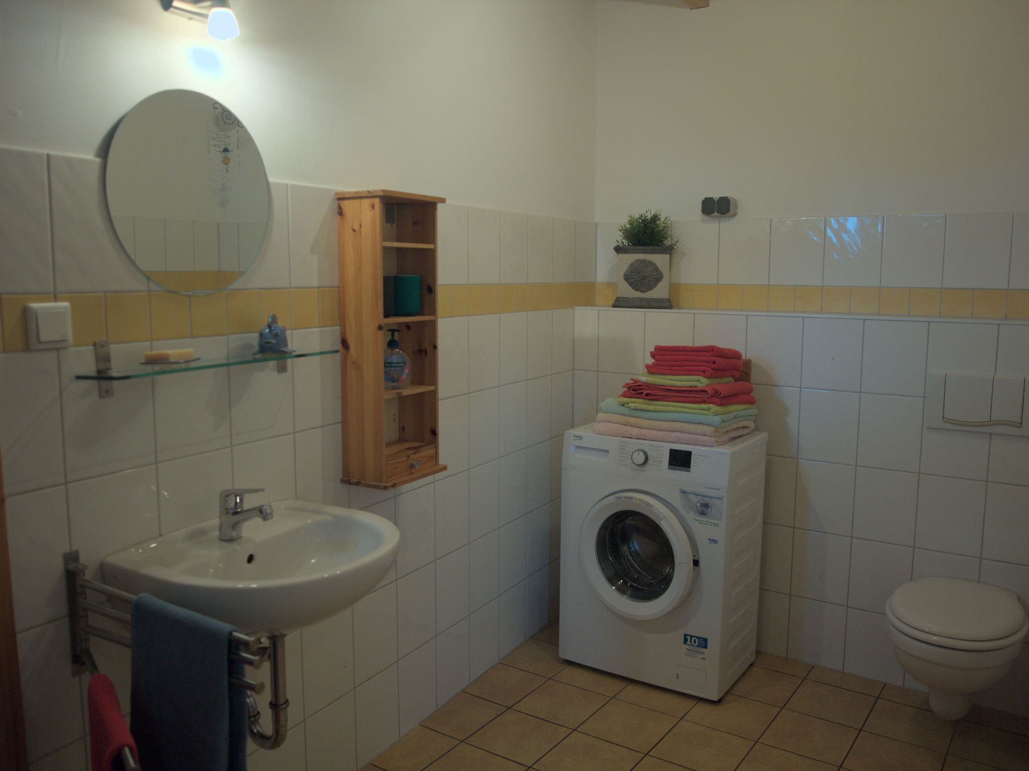Une petite salle de bain comprenant un lavabo, un miroir, un meuble en bois, des serviettes pliées, un lave-linge, des toilettes et une petite plante en pot.
