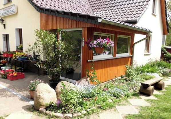 Ein kleines Holzgebäude mit Ziegeldach, einem Blumentopf am Fenster und einem Garten mit verschiedenen Pflanzen und Blumen.