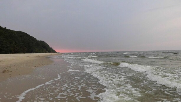 A beach with waves approaching the shore, a forested hill in the background, and a sunset sky. - Schiemann by Interhome (Karnin)