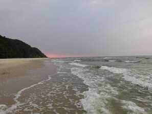 Ein Strand mit ankommenden Wellen, ein bewaldeter Hügel im Hintergrund und ein Sonnenuntergangshimmel.