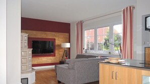 Ein Wohnzimmer mit Flachbildfernseher an einer Holzwand, einem grauen Sofa, einem kleinen Tisch mit Lampe und einem fenster mit Vorhängen.