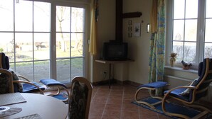 Una sala de estar con televisor, dos mecedoras, una mesa de comedor y ventanas amplias.